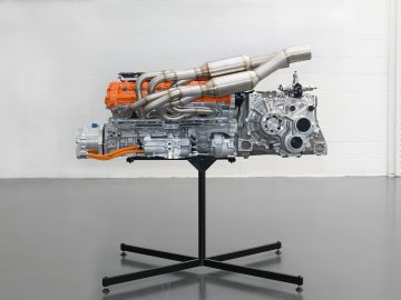 Een complexe hybride automotor tentoongesteld op een standaard in een schone werkplaats, met metalen componenten en oranje kabels, ontworpen door Gordon Murray Automotive T.50.