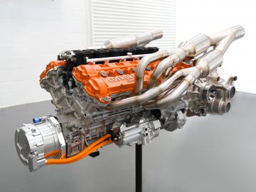 Een complexe Gordon Murray Automotive T.50-motor met zichtbare metalen buizen en een oranje kap, tentoongesteld in een schone, witte kamer.