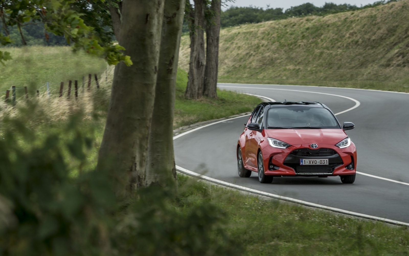 Een Toyota Yaris rijdt op een bochtige weg omzoomd met bomen en hekken onder een bewolkte hemel.