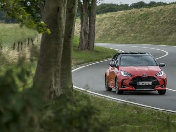 Een Toyota Yaris rijdt op een bochtige weg omzoomd met bomen en hekken onder een bewolkte hemel.