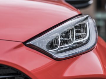 Close-up van de rode koplamp en een deel van de motorkap van een Toyota Yaris, met de nadruk op het gedetailleerde ontwerp en de textuur.