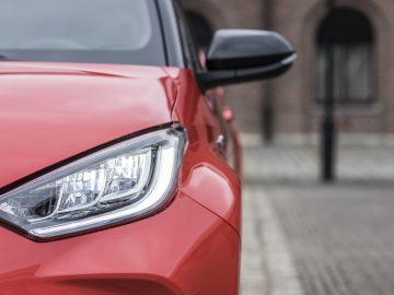 Close-up van de koplamp en het voorste gedeelte van een rode Toyota Yaris met een wazig bakstenen gebouw op de achtergrond.