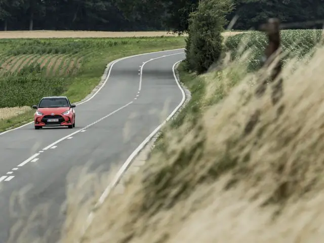 Een rode Toyota Yaris die over een bochtige weg rijdt te midden van weelderige groene velden.