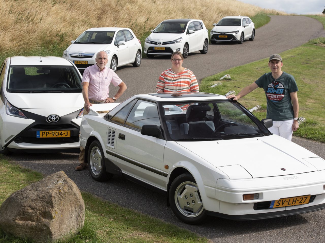Drie mensen staan naast vier verschillende Toyota-voertuigen op een met gras begroeide heuvel en tonen modellen uit verschillende jaren.