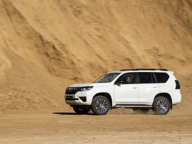 Een witte Toyota Land Cruiser rijdt snel op een zandheuvel onder een heldere hemel.