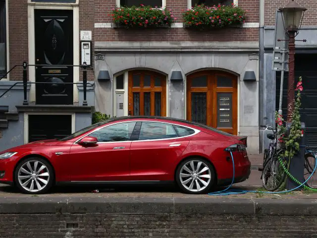 Rode Tesla Model S elektrische auto opladend in een straat vol oude gebouwen, met een fiets vlakbij geparkeerd en Tesla-rijders die een dating-app gebruiken.
