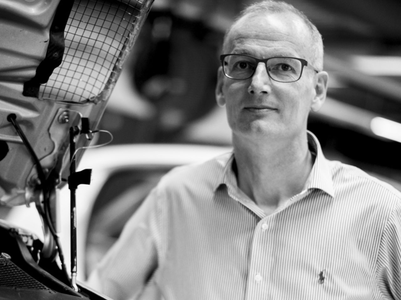 Een man van middelbare leeftijd met een bril staat in een garage, met op de achtergrond een open Peugeot Sport Engineered-autokap; afbeelding in zwart-wit.