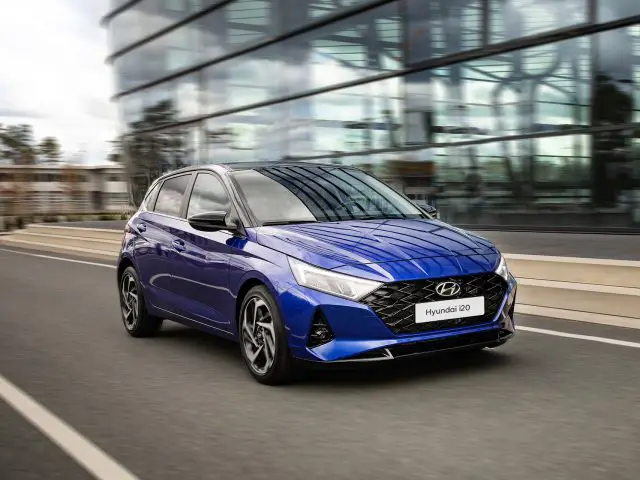 Een blauwe Hyundai i20-auto die over een weg rijdt met moderne glazen gebouwen op de achtergrond.