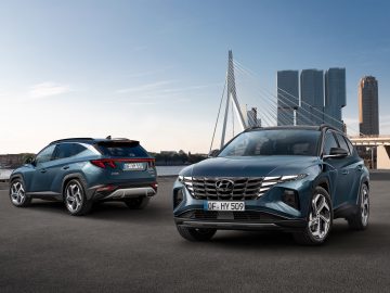 Twee moderne SUV's van Hyundai Tucson geparkeerd aan een promenade aan het water, met een brug en wolkenkrabbers op de achtergrond.