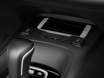 De smartphone van een Suzuki Swace, geplaatst in de draadloze oplaadsleuf van de auto, omgeven door bedieningsknoppen en een versnellingspook.