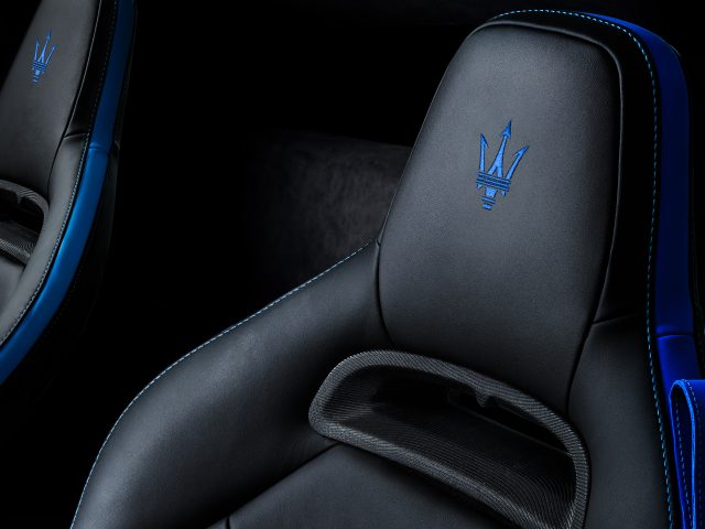 Close-up van luxe Maserati MC20-autostoelen met blauw stiksel en geborduurd embleem op de hoofdsteunen, tegen een donker interieur.