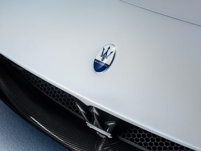 Close-up van een Maserati MC20-logo op de motorkap van een witte auto, met het iconische drietandembleem in een ovale blauwe badge.