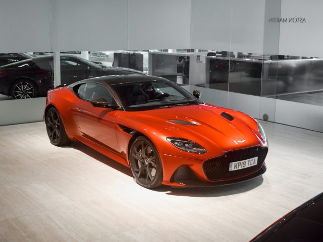 Een oranje Aston Martin DBS Superleggera geparkeerd in een showroom, onder felle lichten, met een Max Verstappen-logo op de muur erachter.