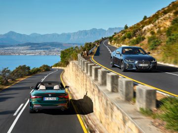 Twee auto's, een BMW 4 Serie Cabrio en een coupé, rijden over een kustweg met een helderblauwe lucht en de oceaan aan de ene kant en bergen in de verte.