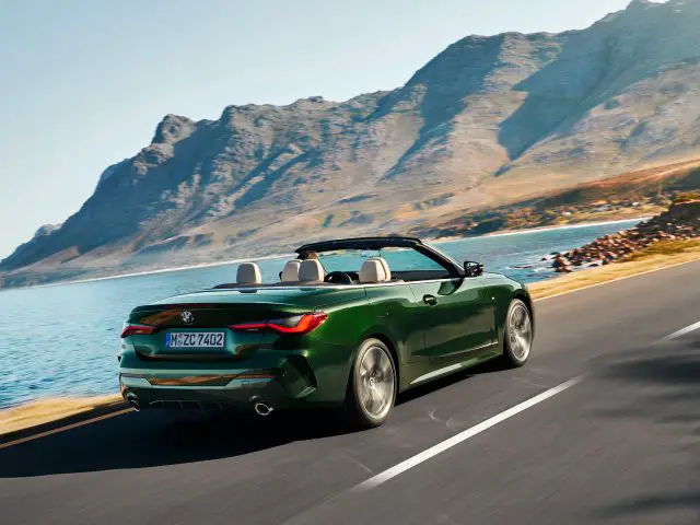Een groene BMW 4 Serie Cabrio rijdt op een zonnige dag langs een kustweg met bergen op de achtergrond.
