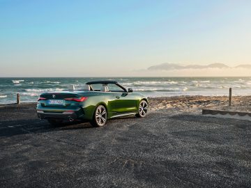 BMW 4 Serie Cabrio geparkeerd op een kustweg met oceaangolven en bergen op de achtergrond bij daglicht.