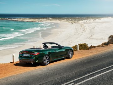 Een groene BMW 4 Serie Cabrio geparkeerd naast een schilderachtige kustweg met turquoise golven die onder een heldere hemel op een zandstrand aanspoelen.