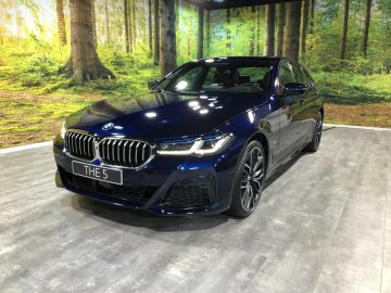 Een blauwe BMW 5 Serie sedan tentoongesteld met een weelderige bosachtergrond tijdens een overdekte autoshow.