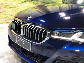 Close-up van een blauwe BMW 5 Serie-auto, gericht op de grille en gedeeltelijk zicht op de koplamp, met een kentekenplaat met de tekst "de 5".