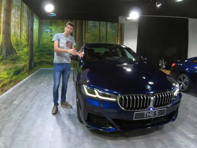 Een man die naast een blauwe BMW 5 Serie sedan in een showroom staat, gebarend naar de auto, met een bosachtige achtergrond.