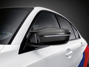 Close-up van de zijspiegel van een BMW M3 Sedan met koolstofvezeldetails op een donkere achtergrond.