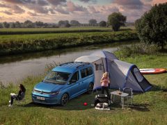 Twee vrouwen kamperen naast een Volkswagen Caddy California en een tent bij een rivier, de een zit bij een stoel en de ander zet een grill op, in een grasveld met kajaks in de buurt.