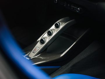 Close-up van de middenconsole van een Ferrari Omologata met drie zilveren knoppen, koolstofvezeldetails en lederen stoelen.
