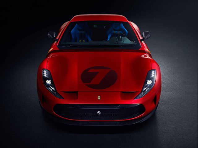 Een rode Ferrari Omologata-sportwagen met een prominent wit nummer 7 op de motorkap, gezien vanuit een hoge hoek aan de voorkant tegen een donkere achtergrond.