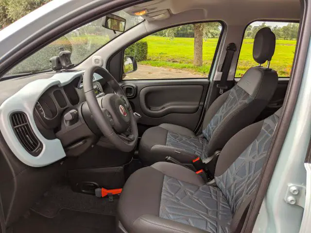 Interieur van een Fiat Panda met de bestuurdersstoel, het stuur en het dashboard, met een open deur en uitzicht op een groen landschap buiten.