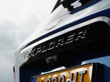 Close-up van de achterkant van een blauwe Ford Explorer PHEV met een 'Explorer'-badge en een Europees kenteken, onder een bewolkte hemel.