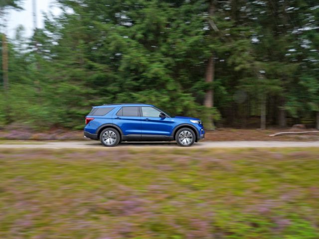 Een blauwe Ford Explorer PHEV die snel over een met bos omzoomde weg rijdt, waarbij bewegingsonscherpte de snelheid van het voertuig benadrukt.