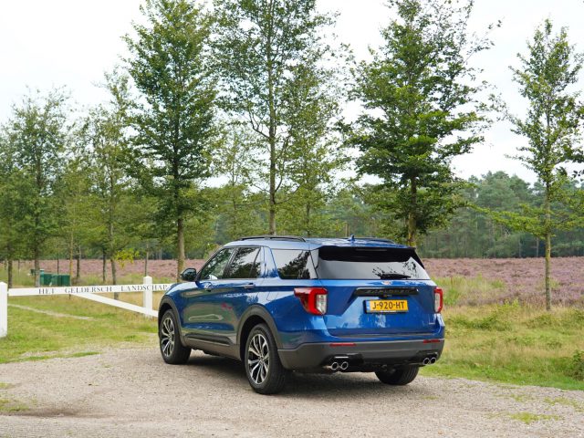 Blauwe Ford Explorer PHEV geparkeerd op een onverharde weg nabij een bos, met een veld met paarse bloemen op de achtergrond.