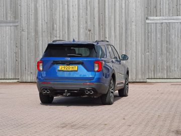 Een blauwe Ford Explorer PHEV geparkeerd voor een houten schuur, met het achteraanzicht van het voertuig met zichtbaar kenteken "j920-ht".