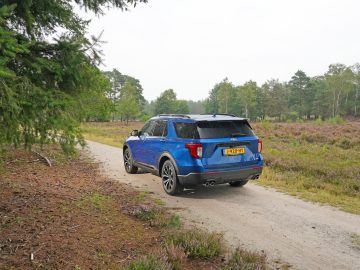 Een blauwe Ford Explorer PHEV geparkeerd op een onverharde weg naast een bosrijk gebied en open heide onder een heldere hemel.