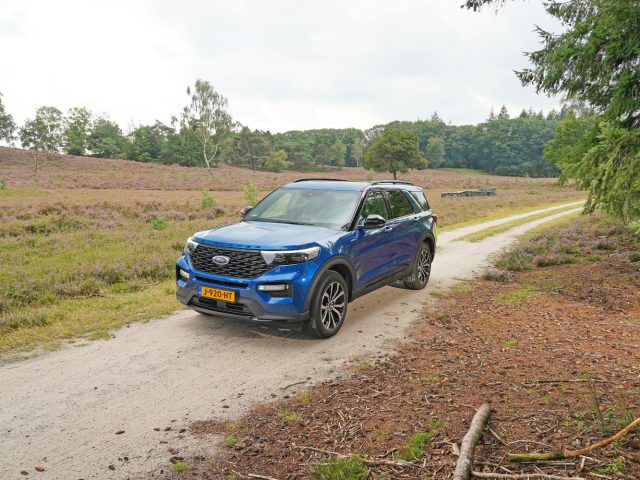 Een blauwe Ford Explorer PHEV geparkeerd op een onverhard pad in een heidegebied met verspreide bomen en een bewolkte lucht.