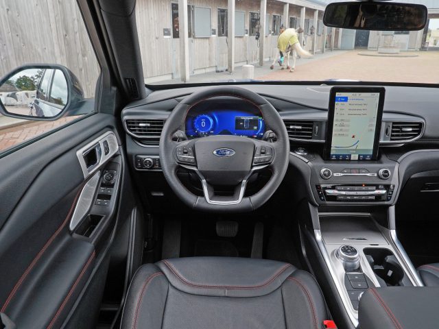 Binnenaanzicht van een moderne Ford Explorer PHEV met de nadruk op het dashboard, het stuur en het multimediasysteem, geparkeerd met zicht op mensen buiten.