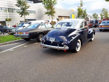 Oldtimers rijden op straat tijdens de Saturday Night Cruise 2020, waaronder een donkerblauwe klassieke sedan met zichtbare kentekenplaat, op een autoshow.