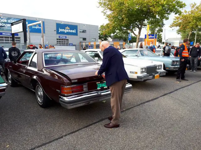 Een oudere man onderzoekt een klassieke kastanjebruine auto tijdens de 'Saturday Night Cruise 2020', een autoshow in de open lucht met andere vintage voertuigen en toeschouwers op de achtergrond.