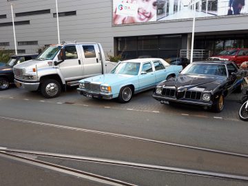 Drie oldtimers geparkeerd op straat, met een wit busje, een lichtblauwe sedan en een zwarte coupé, buiten een modern gebouw tijdens de Saturday Night Cruise 2020.