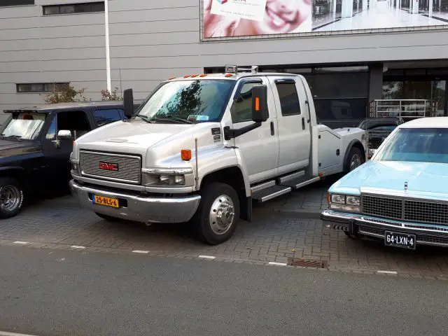 Een grote witte GMC-truck geparkeerd tussen een zwarte pick-up en een blauwe vintage auto in een stadsstraat tijdens de Saturday Night Cruise 2020.