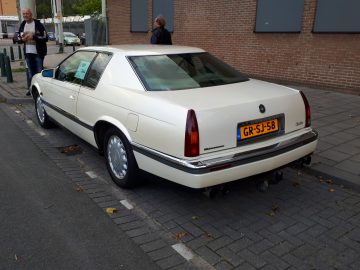Een beige Cadillac Coupe Deville geparkeerd op straat tijdens de Saturday Night Cruise 2020 met een man op de achtergrond.