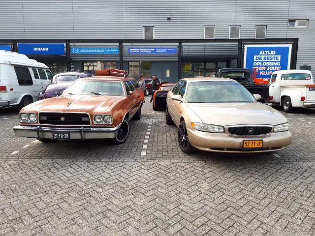 Twee oldtimers, een bruine stationwagen en een zilveren sedan, geparkeerd voor een gebouw met Nederlandse bewegwijzering tijdens de Saturday Night Cruise 2020.