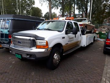 Een wit-gouden sleepwagen geparkeerd tijdens de Saturday Night Cruise 2020, met een oranje vintage auto geladen op de laadvloer, omgeven door bomen en andere voertuigen.