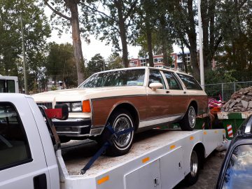 Een vintage bruin-witte sedan wordt vervoerd op een sleepwagen met dieplader tijdens de Saturday Night Cruise 2020 in een stedelijk gebied met bomen en geparkeerde auto's.