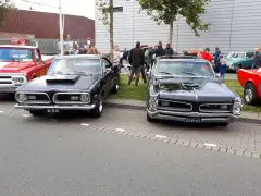 Twee klassieke muscle car's, een zwarte Dodge Charger en een zilveren Pontiac GTO, geparkeerd op de Saturday Night Cruise 2020 met toeschouwers op de achtergrond.