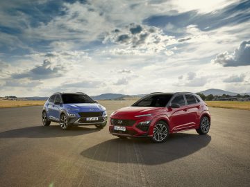 Twee moderne auto's, een zilveren Hyundai Kona en een rode, geparkeerd op een open weg te midden van een uitgestrekt landschap onder een bewolkte hemel.