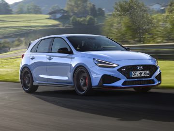 Een blauwe Hyundai i30 N die op een zonnige dag over een weg rijdt met een groen, heuvelachtig landschap op de achtergrond.