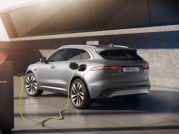 Een zilveren Jaguar F-Pace elektrische SUV staat geparkeerd en wordt opgeladen bij een laadstation buiten, met moderne architectuur op de achtergrond.