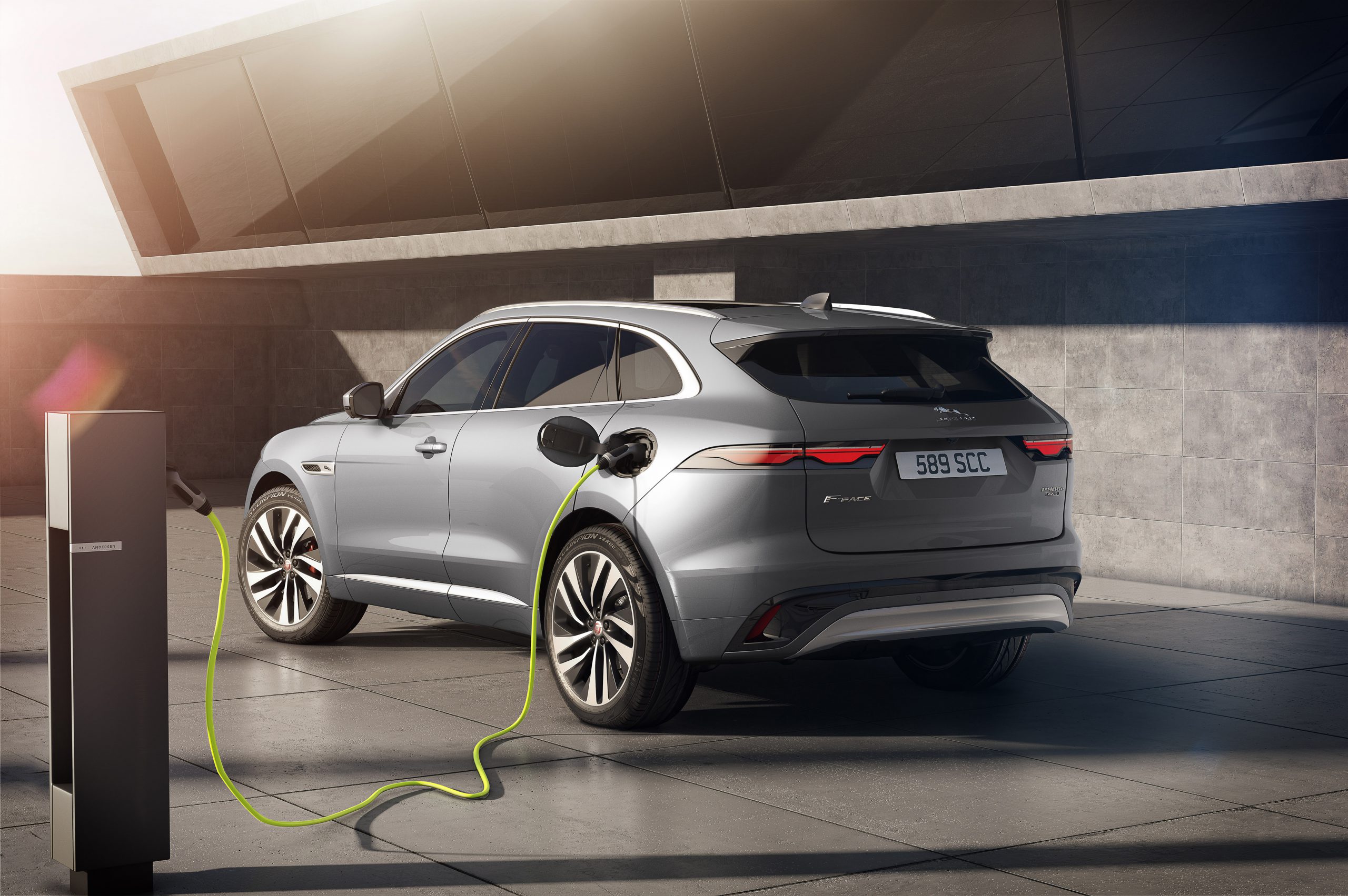 Een zilveren Jaguar F-Pace elektrische SUV staat geparkeerd en wordt opgeladen bij een laadstation buiten, met moderne architectuur op de achtergrond.