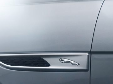 Close-up van de zijventilatie van een zilveren Jaguar F-Pace met een chromen jaguar-embleem.